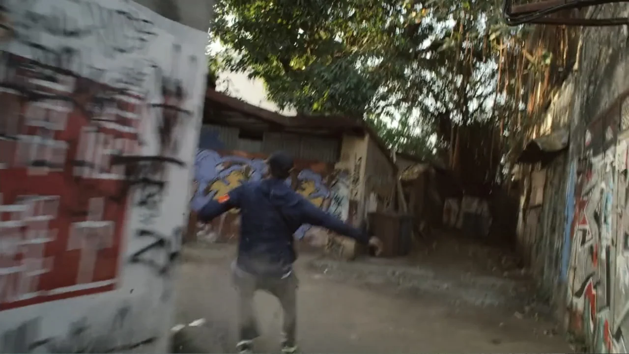 Unknown #1 movie still: fullbody — A narrow alleyway features concrete walls covered in faded graffiti and overhanging leafy trees. A man in a dark…