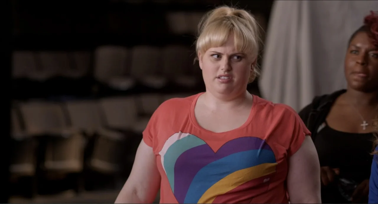 Pitch Perfect (2012) movie still: medium — A dimly lit auditorium interior features rows of empty seats in the soft background. A young woman in a…