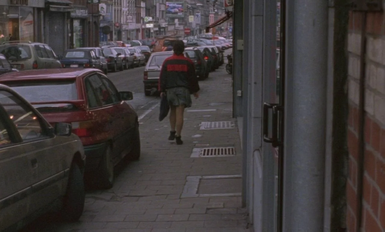 Rosetta (1999) movie still: fullbody — A dense urban sidewalk borders a row of parked cars and shop storefronts. A person walks away down the…