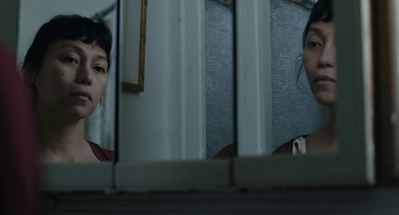Lingua Franca (2019) movie still: A woman stares into her reflection in a framed mirror, her contemplative gaze dominating the left side of the…