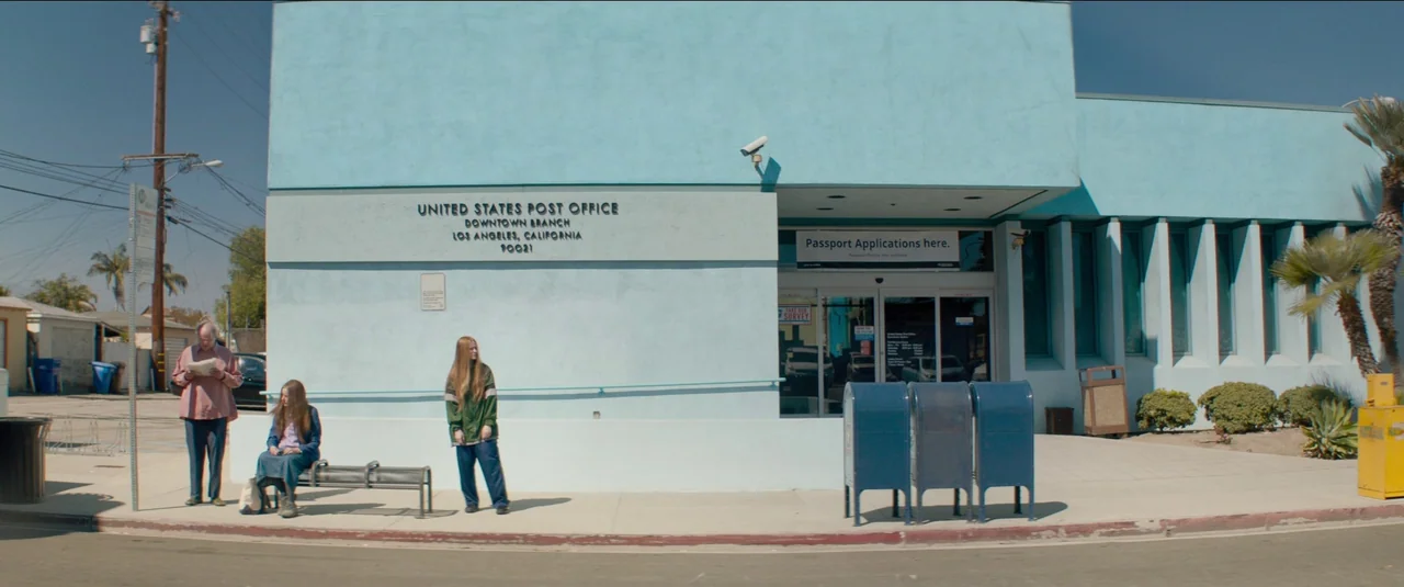 Kajillionaire (2020) movie still: This wide, eye-level shot captures a mundane yet striking street scene outside a pastel blue United States Post…