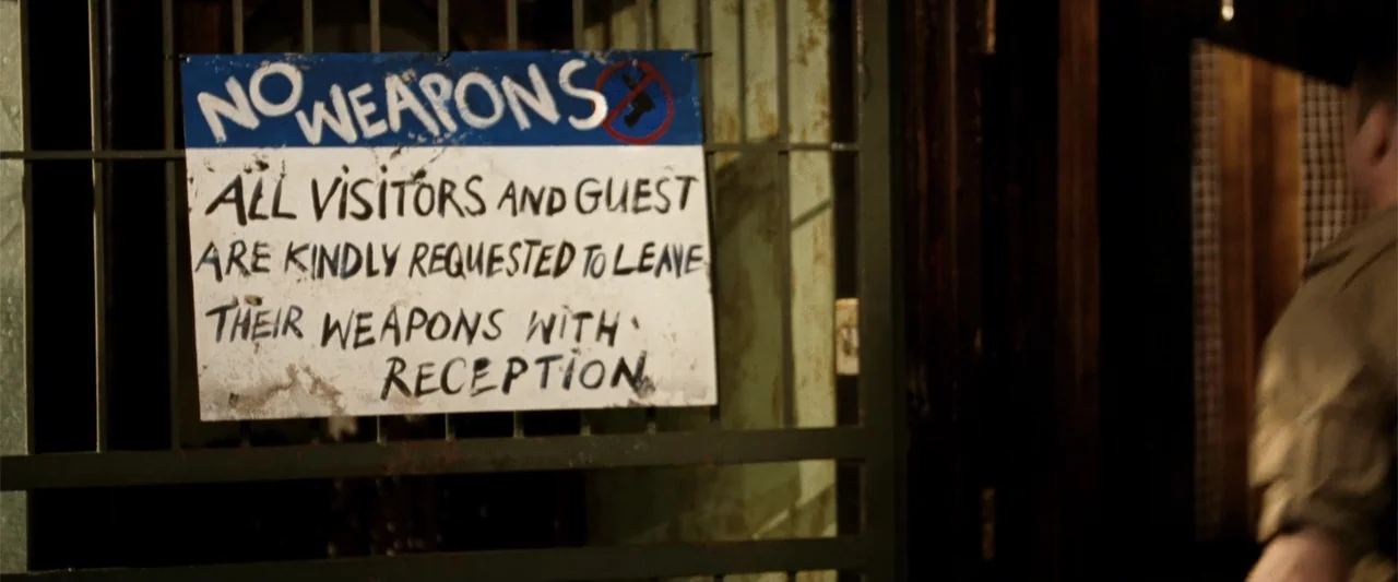 Hector and the Search for Happiness (2014) movie still: medium — A sign hangs on a metal gate in a reception area with text about weapon policy. A…