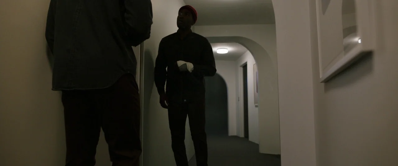 Candyman (2021) movie still: medium — A dimly lit modern apartment hallway features arched doorways and muted white walls. Two men stand facing each…
