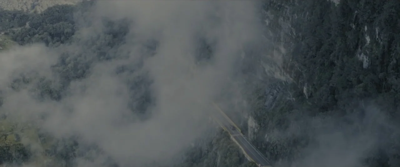 Quicksand (2023) movie still: establishing — A winding asphalt road cuts through a steep, tree-covered mountainside under thick heavy fog. A small…