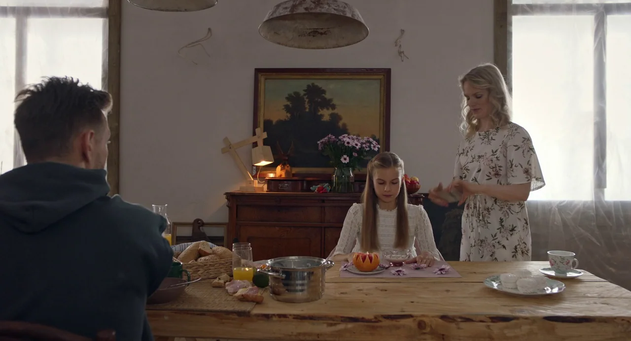 Hatching (2022) movie still: medium — A rustic dining room features a long wooden table with plates and food items. A woman and child face the…