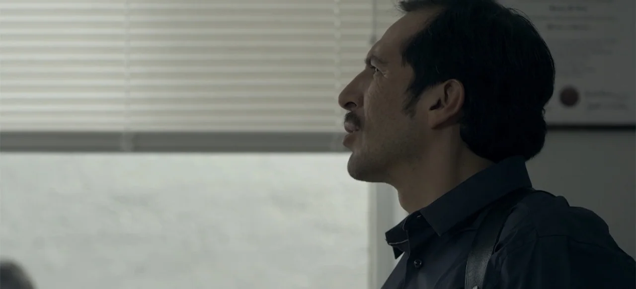 About the Deaths (2016) movie still: closeup — An interior office space with horizontal window blinds and a document on the wall. A man in his…
