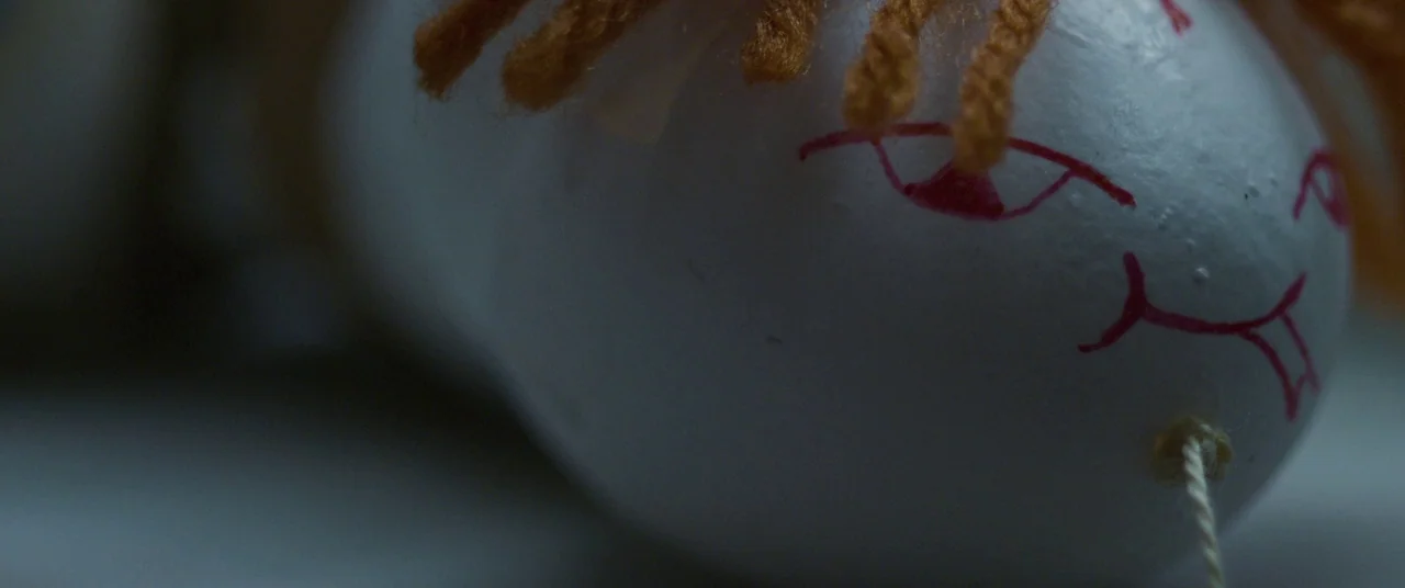 Room (2015) movie still: closeup — A close-up view of a white egg with a hand-drawn red face. A small bunch of orange yarn represents hair on top of…
