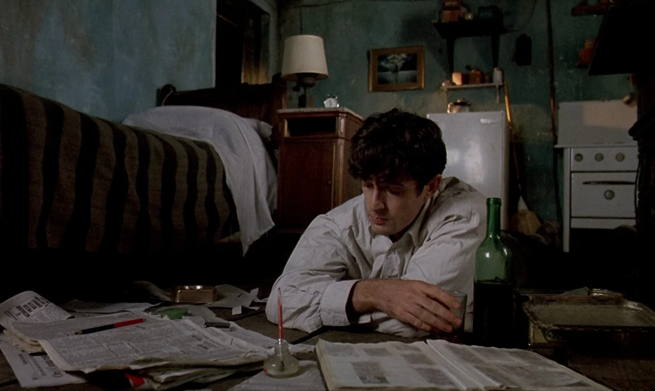 Dellamorte Dellamore (1994) movie still: medium — A cramped bedroom with peeling blue walls, a bed, and a wooden nightstand. A man lies on the floor…