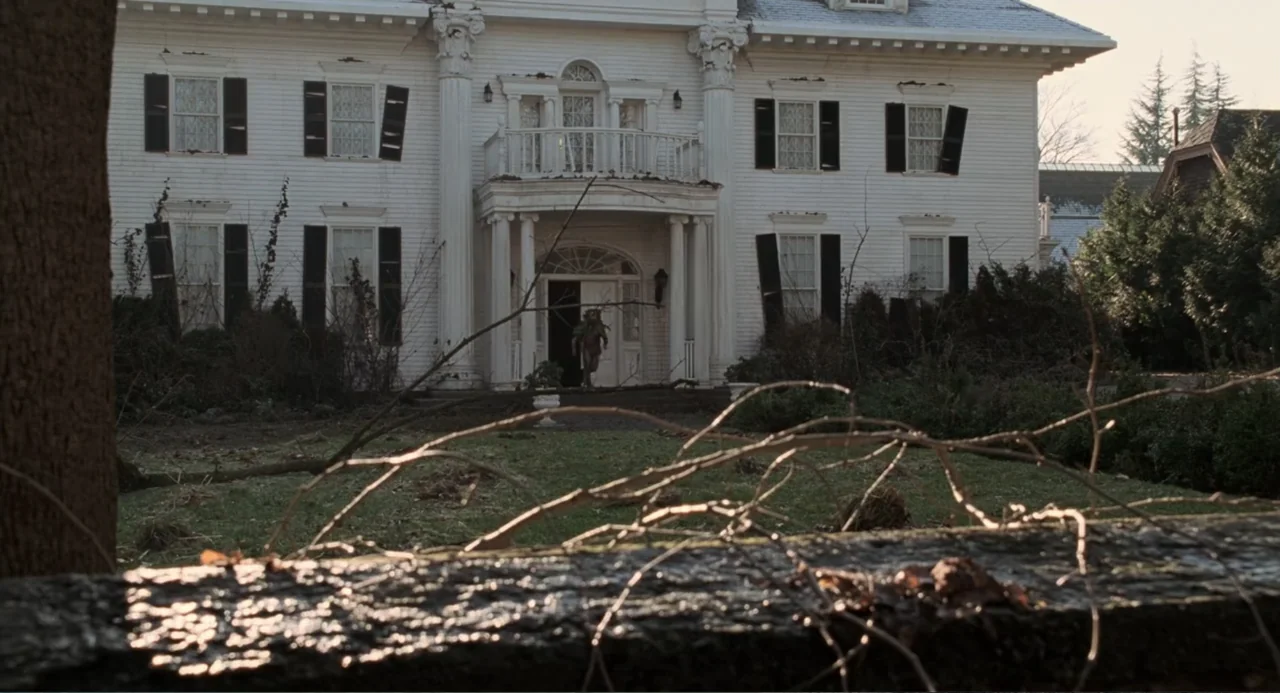 Jumanji (1995) movie still: establishing — A large two-story white neoclassical mansion features a prominent porch and black window shutters. The…