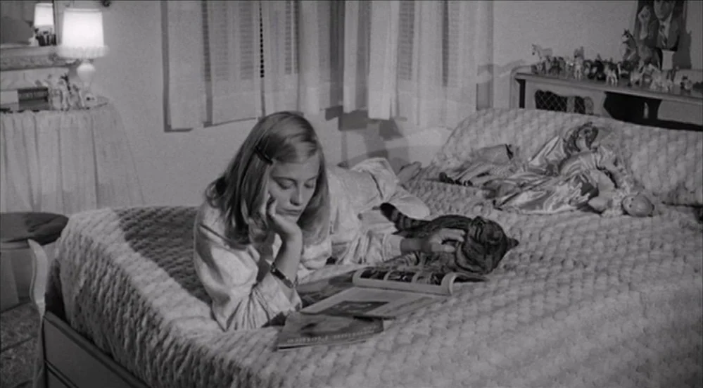 The Last Picture Show (1971) movie still: medium — A teenage girl reclines on a textured bedspread in a room with draped windows. She reads a…
