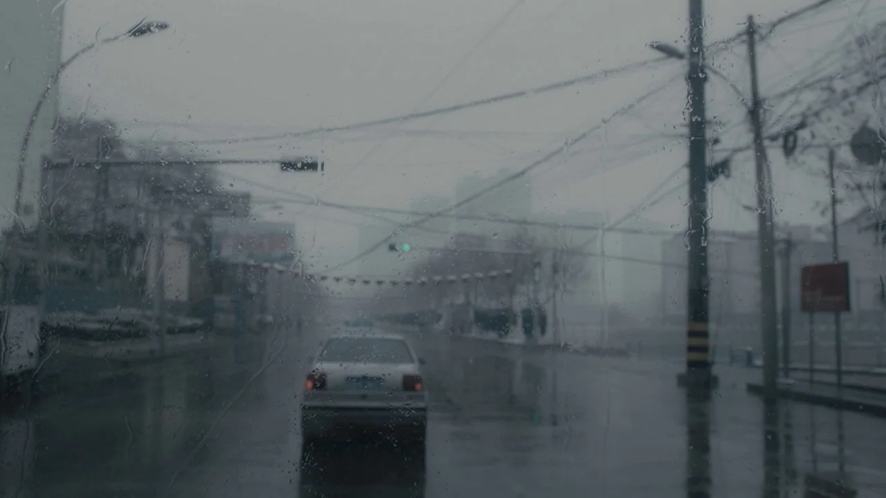 An Elephant Sitting Still (2018) movie still: wide — A rain-covered window looks out onto a gray, misty city street at intersection. A light-colored…