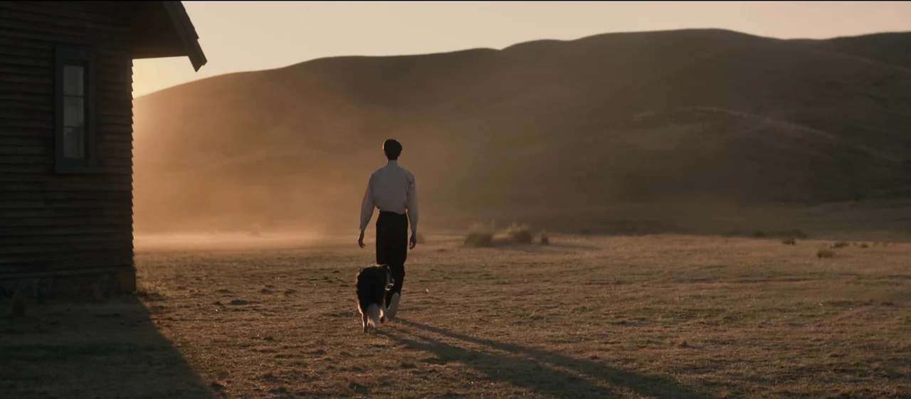 The Power of the Dog (2021) movie still: wide — A desolate open field stretches toward rounded hills under a bright, low-hanging sun. A thin man…