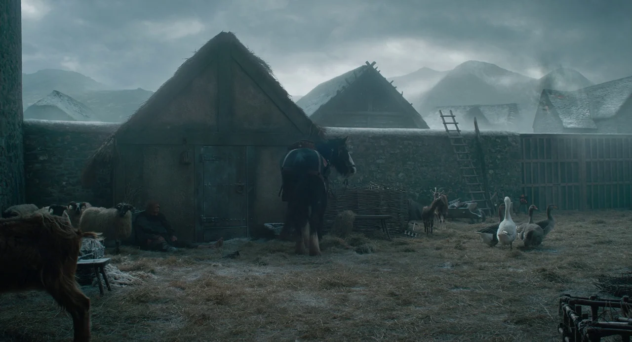 The Green Knight (2021) movie still: wide — A frost-covered medieval barnyard features thatched huts against a background of misty mountains. A…