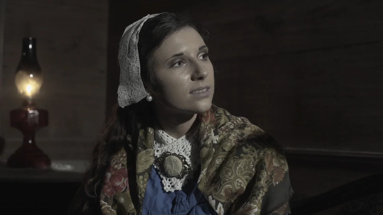The Dalton Gang (2020) movie still: medium — A dark wooden room interior features a woman illuminated by a nearby glowing lamp. She wears a…
