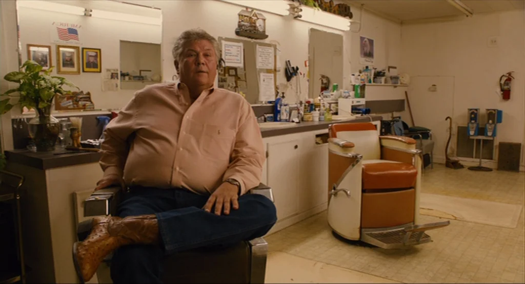 Bernie (2011) movie still: medium — A dated barbershop with vintage vinyl chairs, framed photographs on the wall, and counters. An older man wearing…