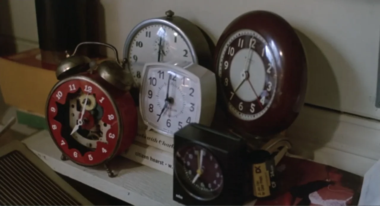Dangerously Close (1986) movie still: closeup — A collection of various analog alarm clocks sits arranged on a white shelf. Several clocks with…