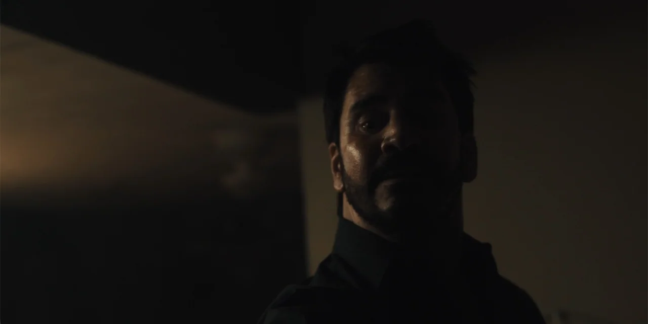 All Roads to Pearla (2019) movie still: closeup — A dimly lit indoor space with dark walls and minimal environmental detail. A man stands facing the…