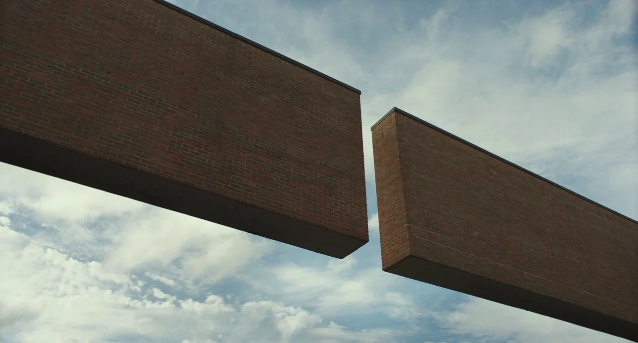 Columbus (2017) movie still: establishing — Two monolithic red brick structures project horizontally into the air against a cloudy sky. The…