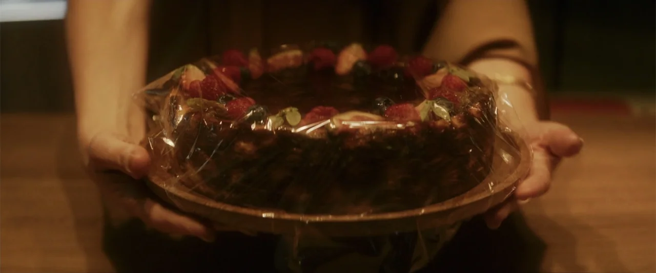 Black Bag (2025) movie still: closeup — A dark indoor setting features a wooden plate held by two hands. The plate holds a dark cake topped with…