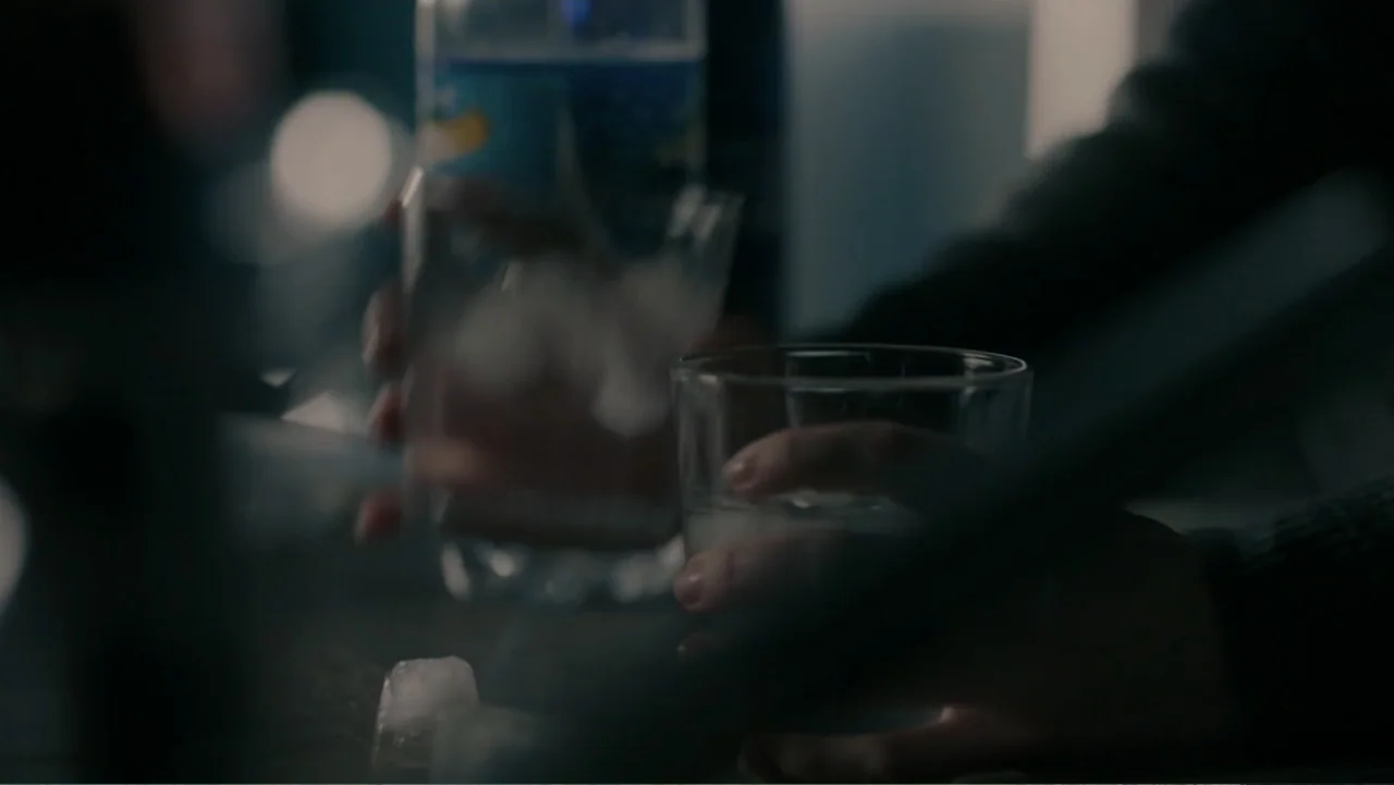 Sightless (2017) movie still: closeup — A dark bar setting features hands holding glass tumblers containing ice and liquid. The blurred background…