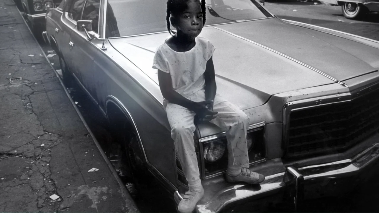 Mary J. Blige's My Life (2021) movie still: fullbody — A young girl sits on the hood of a parked sedan on a city street. The girl looks toward the…
