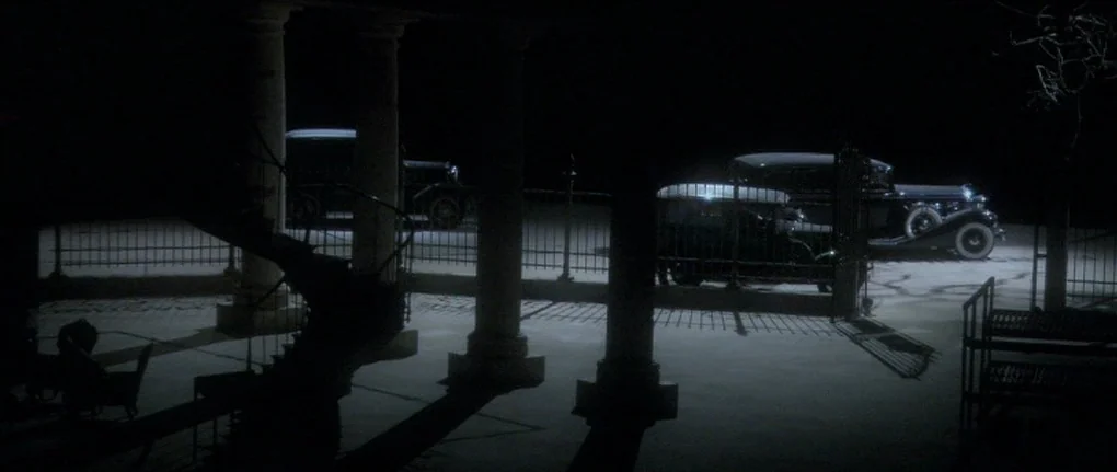 Manderlay (2005) movie still: wide — An exterior night view shows a paved area framed by stone columns and fencing. Two vintage luxury cars are…