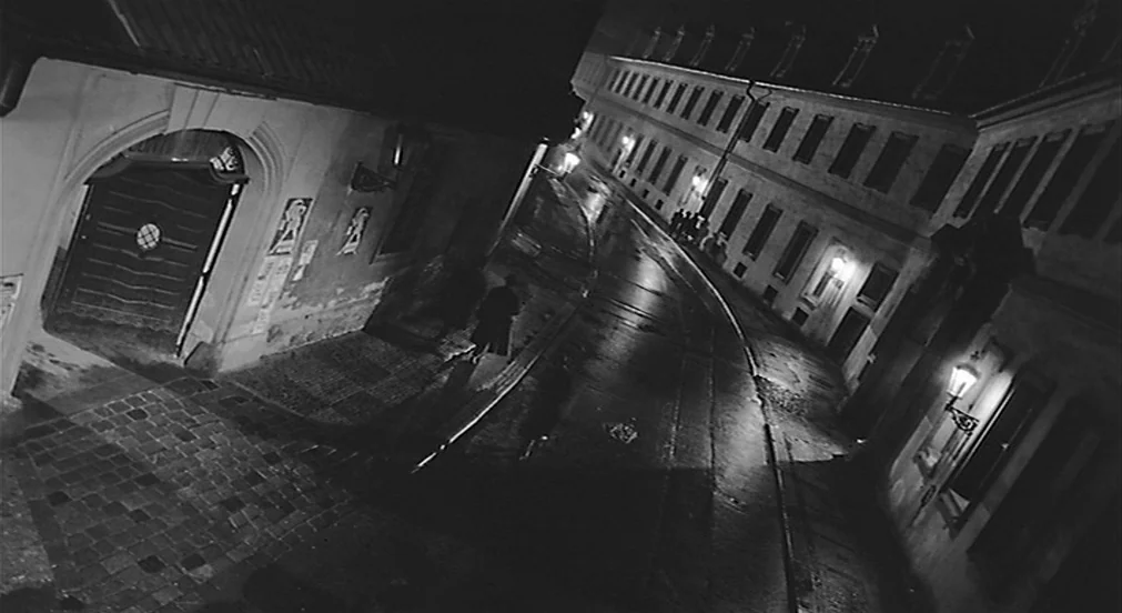 Kafka (1991) movie still: wide — A narrow, wet cobblestone street curves between tall European-style buildings in the dark night. A small group of…