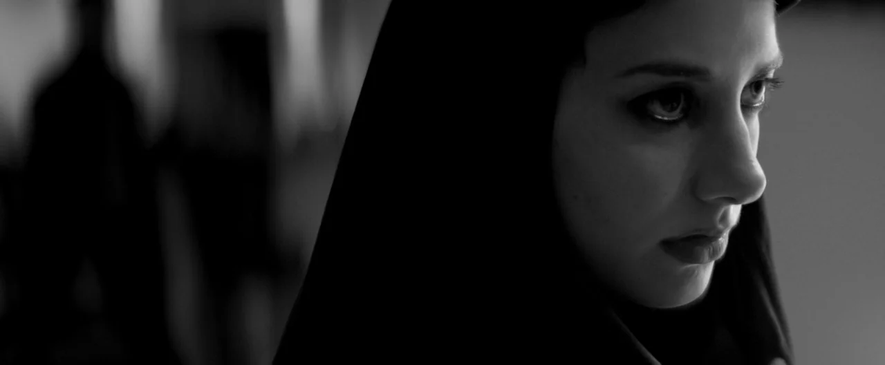 A Girl Walks Home Alone at Night (2014) movie still: closeup — A close-up view shows a young woman in a dark headscarf. She looks toward the side of…