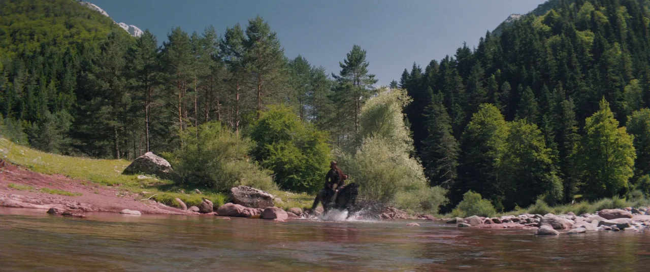 The Sisters Brothers (2018) movie still: wide — A wide, shallow river flows through a dense pine forest with rocky banks. A lone rider on a black…