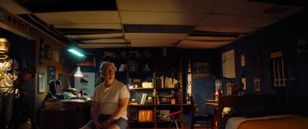 Linoleum (2022) movie still: medium — A basement room with dark blue walls is filled with space-themed models and books. A middle-aged man sits on a…