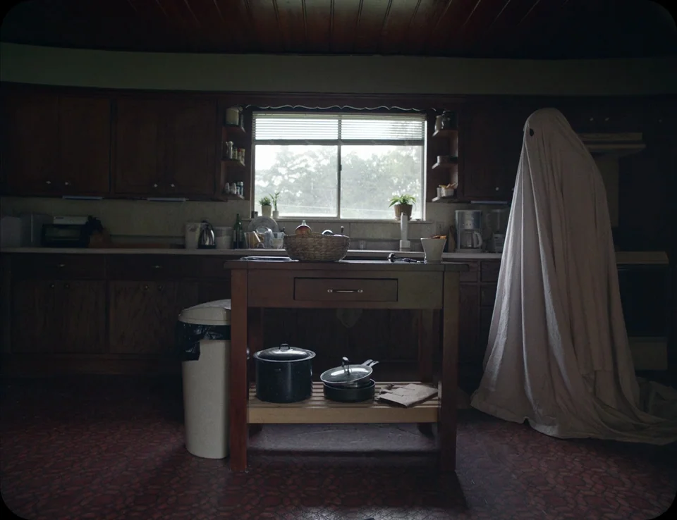 A Ghost Story (2017) movie still: medium — A residential room features dark wooden cabinets, a central island, and patterned flooring. A figure…