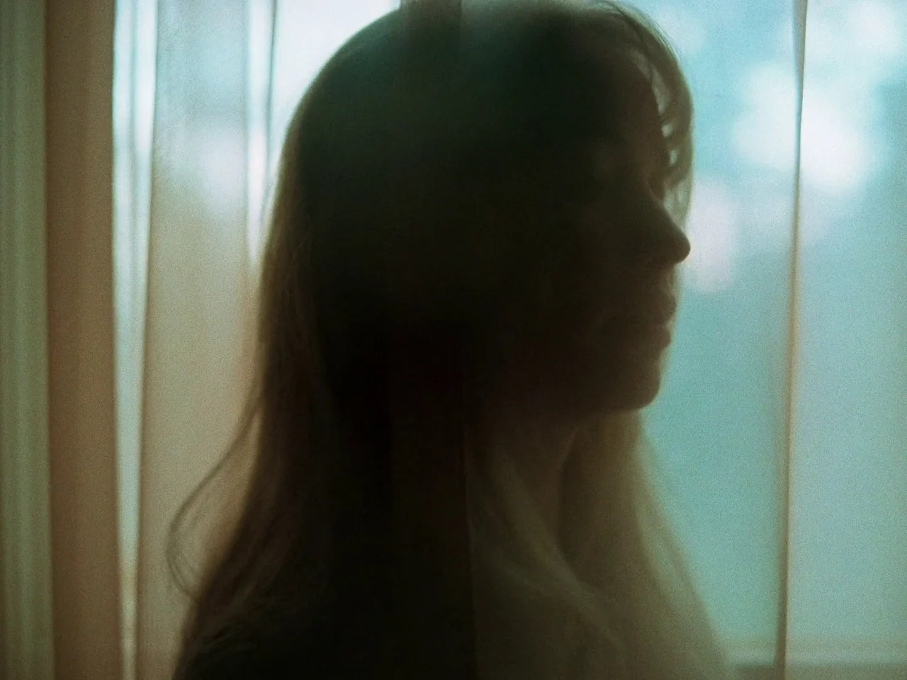 Die My Love (2025) movie still: closeup — A silhouette of a person stands against a bright window covered by thin curtains. Natural daylight washes…