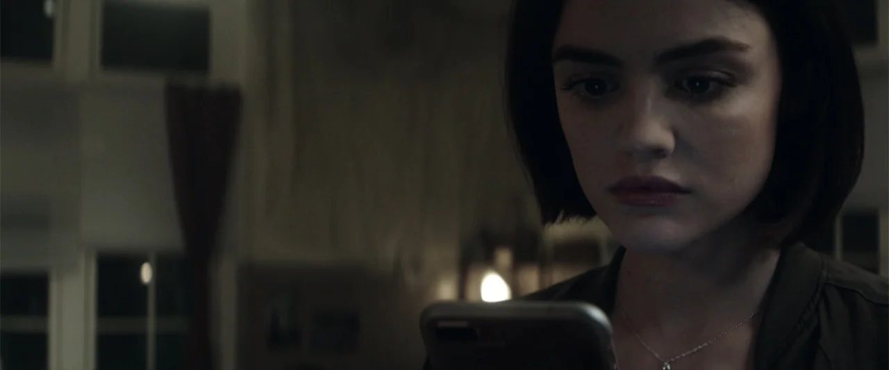 Truth or Dare (2018) movie still: closeup — A dimly lit residential room features a window in the blurred background. A young woman views her…
