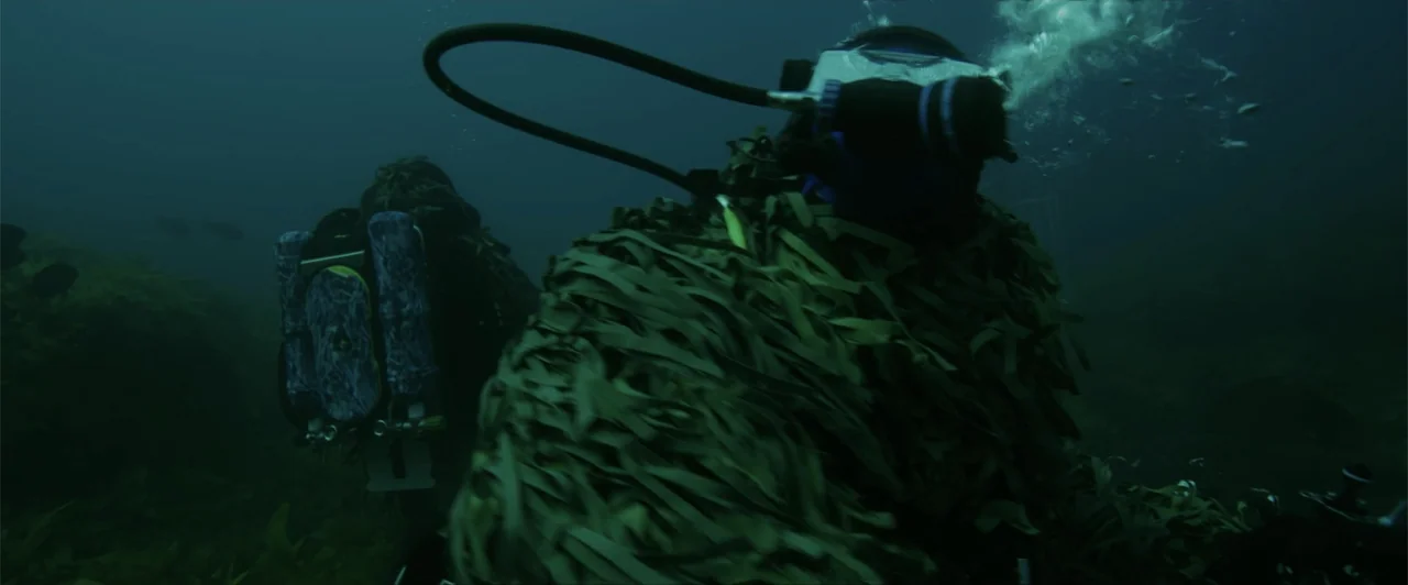 Lair of the Great White (2024) movie still: medium — Two divers in camouflage suits move through murky, dark, deep water. The subjects swim…