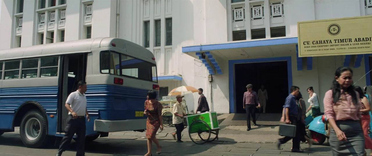 Satan’s Slaves (2017) movie still: wide — An urban street scene featuring a large blue and white bus parked near a building. Several individuals…