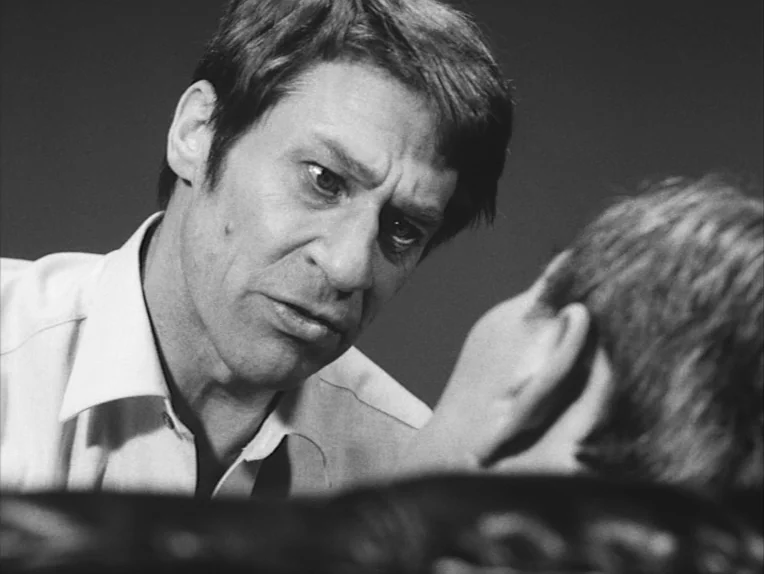 The Rite (1969) movie still: closeup — A close-up shot focuses on a man in a light shirt looking downwards. He appears to be addressing a second…