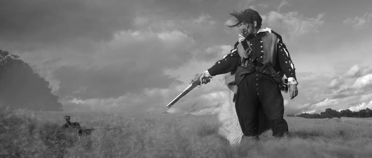 A Field In England (2013) movie still: fullbody — An open grassy field under a cloudy, overcast sky during the day. A bearded man in period clothing…