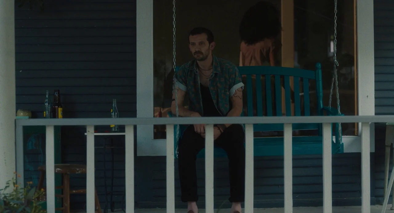 Test Pattern (2019) movie still: medium — A dark blue house porch features a teal hanging swing bench and wooden stool. A man in a patterned shirt…