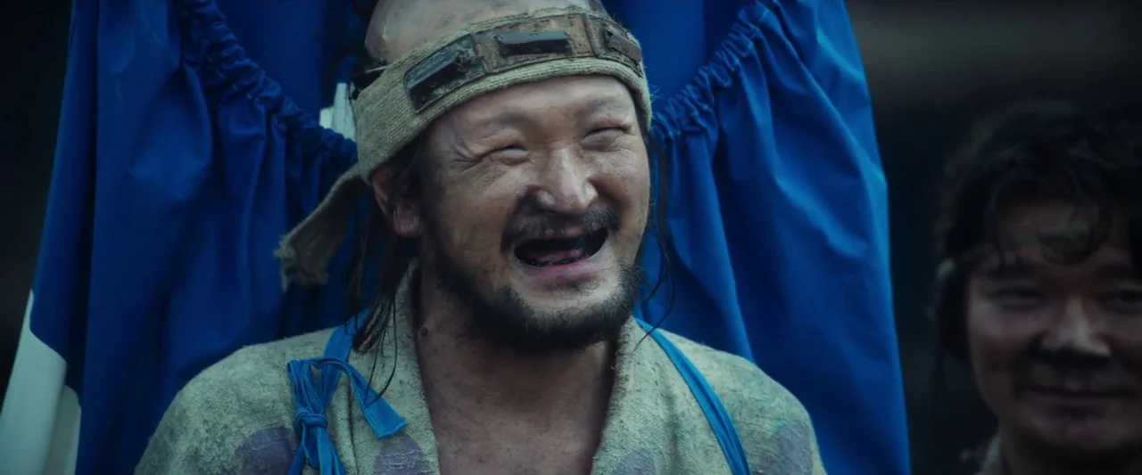 Kubi (2023) movie still: medium — A man with a shaved head and dirty face stands against a blue backdrop. He wears a rough beige garment and laughs…