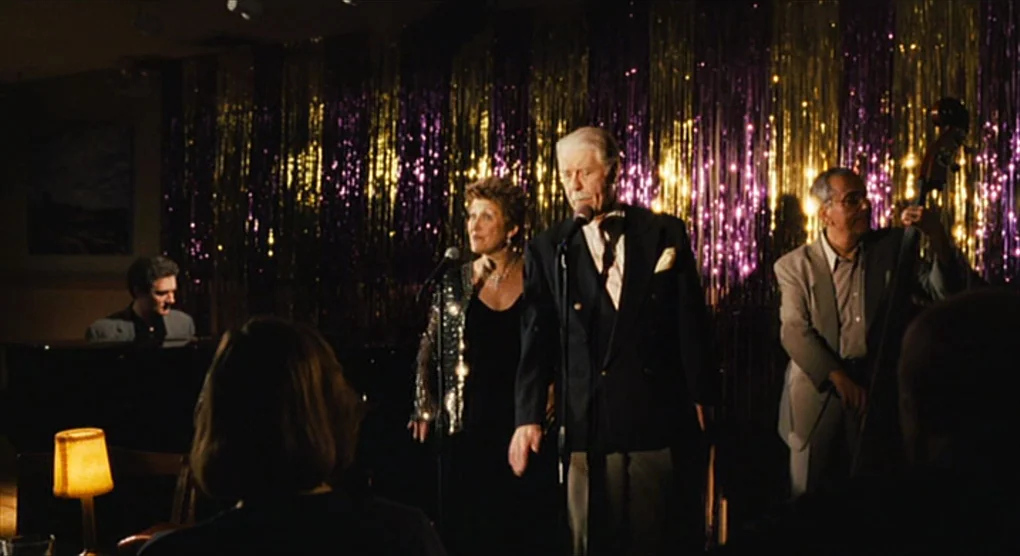 The Savages (2007) movie still: medium — A dimly lit lounge features a stage decorated with vertical gold and purple tinsel. A musical trio stands…
