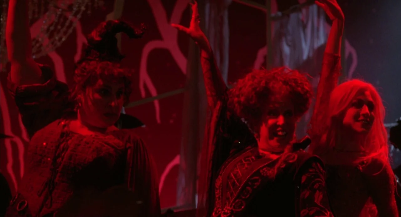 Hocus Pocus (1993) movie still: medium — A dark stage set with red light bathing three women in period costumes. The three women dance and sing with…