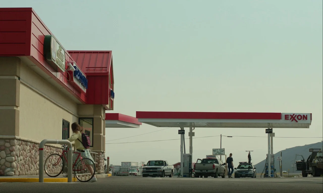 Mickey and the Bear (2019) movie still: wide — A modern gas station with red-and-white architecture under a hazy, pale sky. A person walks a red…