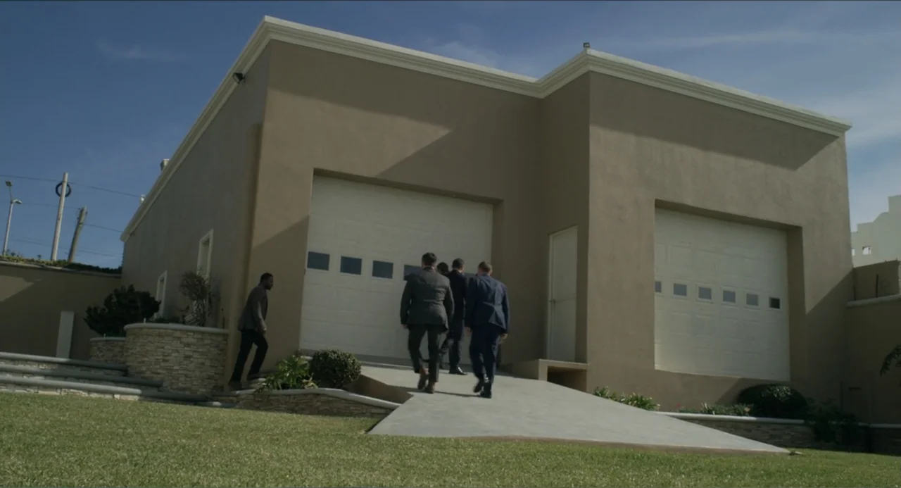 Instantes Capturados (2022) movie still: wide — A modern beige residential house features two white garage doors and a concrete ramp. Four men in…