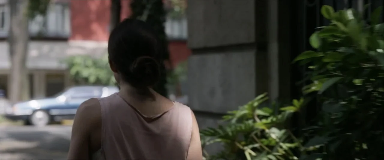Dive (2013) movie still: medium — A residential city sidewalk features a stone building facade and a leafy green shrub. A young woman with her hair…