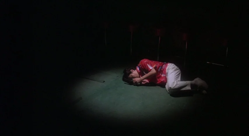 Mishima: A Life in Four Chapters (1985) movie still: wide — A dark, empty floor space features a single circular pool of light. A young man lies on…