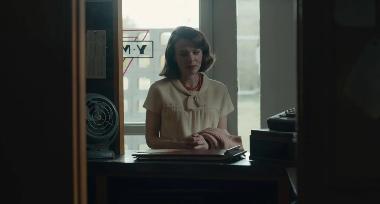 Wildlife (2018) movie still: medium — A woman stands behind a wooden service counter in a mid-century office space. She rests her hands on a…