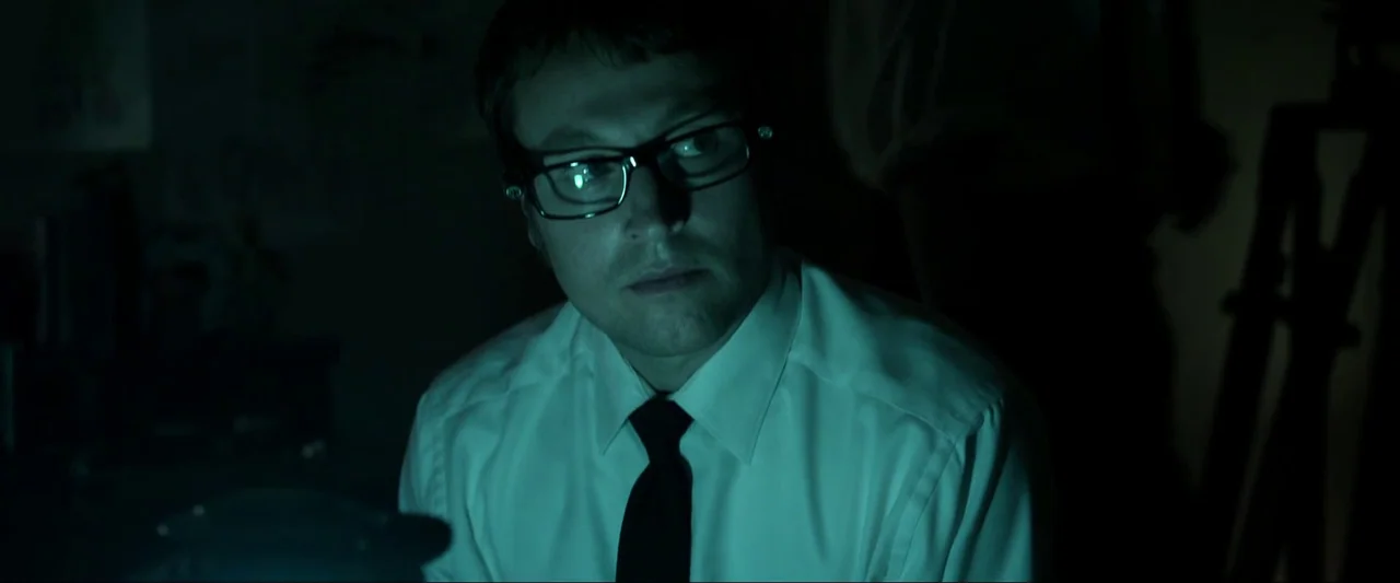 Insidious (2010) movie still: closeup — A dark, dimly lit indoor space with low light levels and high contrast. A man wearing a white shirt and…