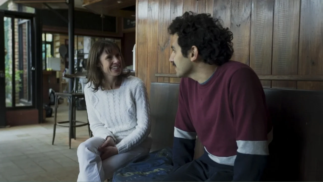 Smog in Your Heart (2022) movie still: medium — A rustic cafe interior features vertical wooden wall paneling and a tiled floor. A woman in a white…