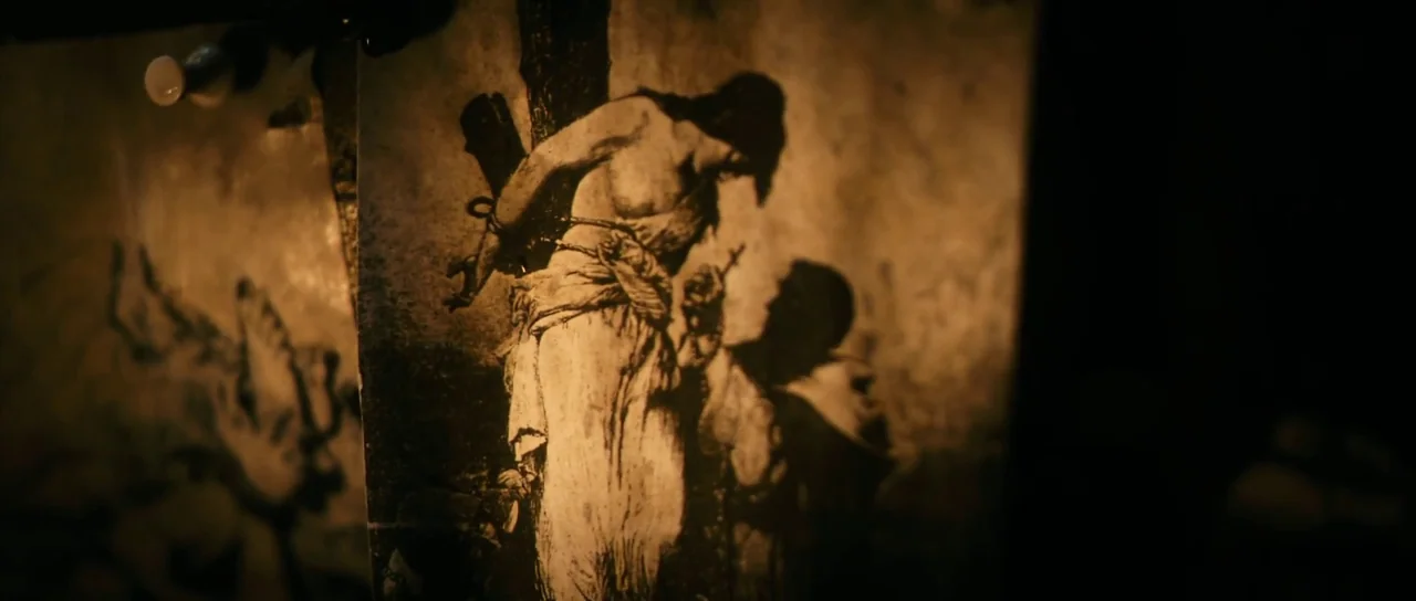 Antichrist (2009) movie still: closeup — A close-up view of a printed image pinned to a dark, unlit wall. The print depicts a figure tied to a post…