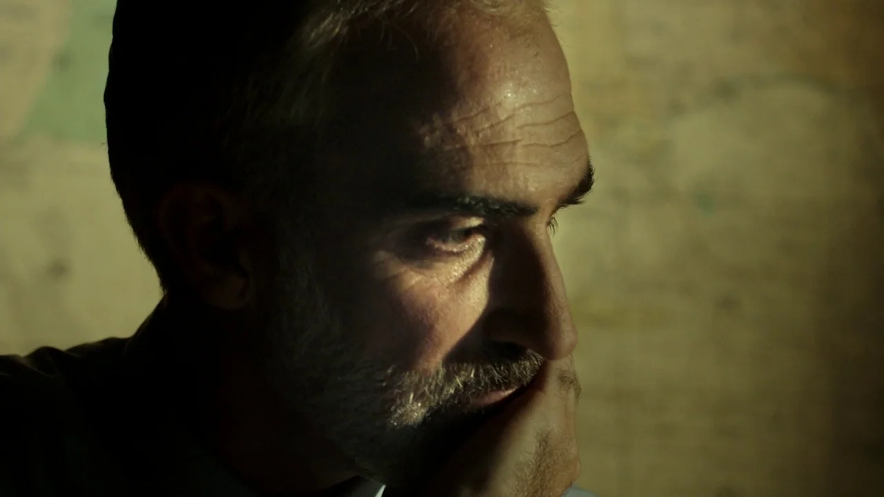The Pact II movie still: closeup — A dimly lit room contains a wall map visible in the blurred background. A middle-aged man with a graying beard…